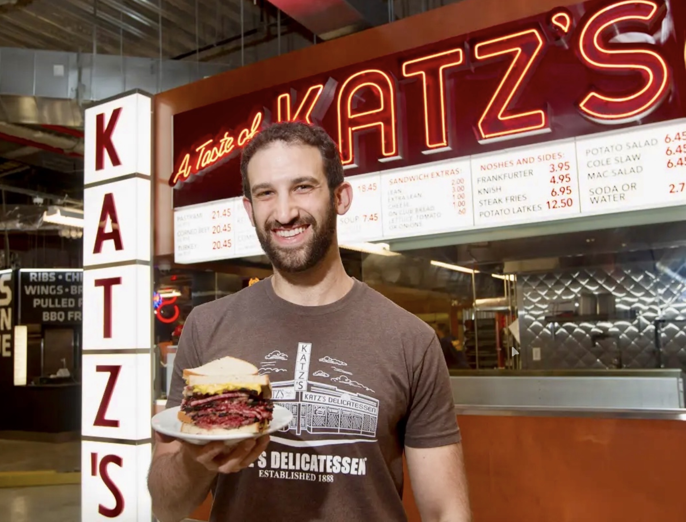 A smiling Katz’s counterman holding a pastrami sandwich in front of the neon A Taste of Katz’s sign