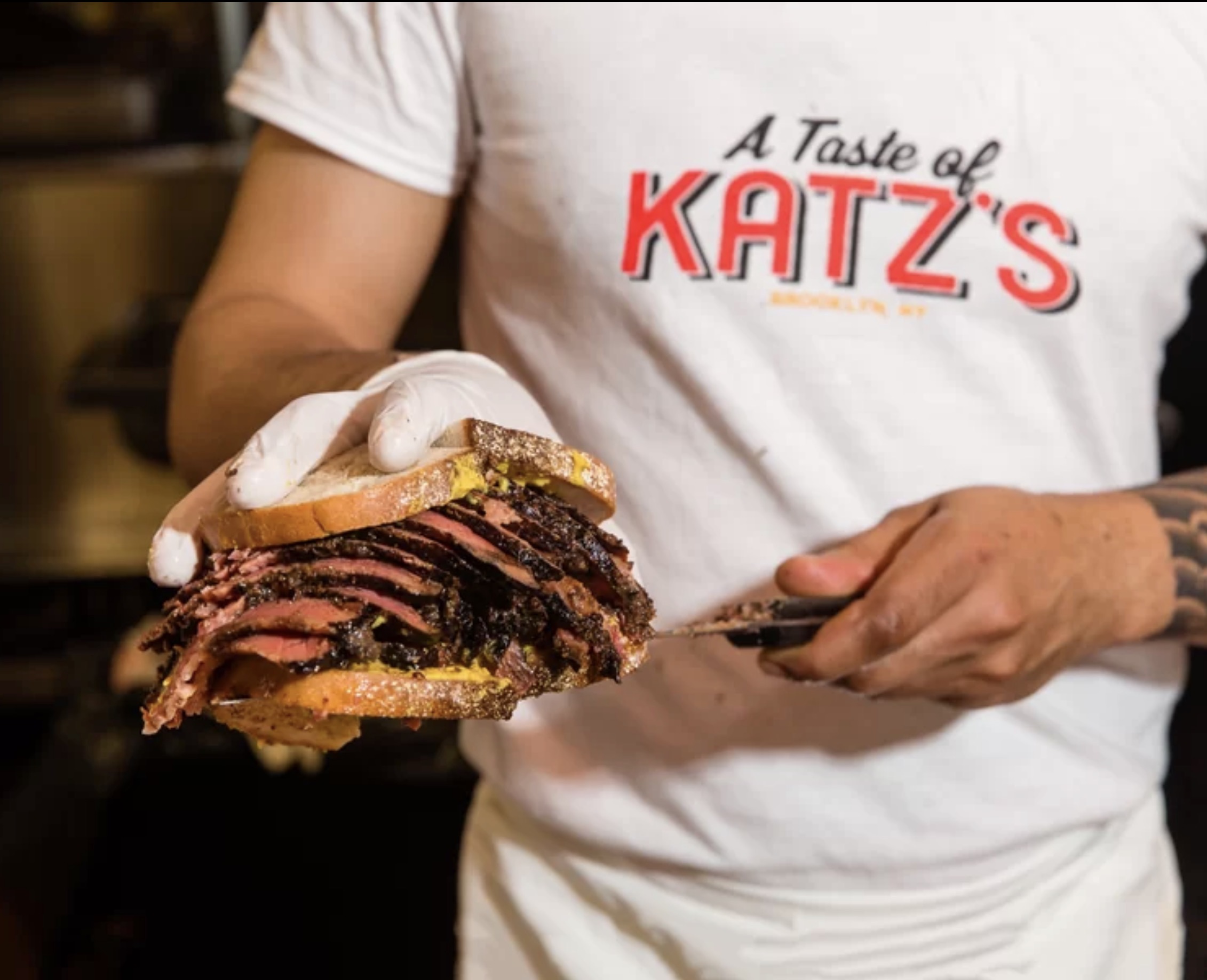 A cook in an A Taste of Katz’s T-shirt holding a hand-carved pastrami sandwich on rye